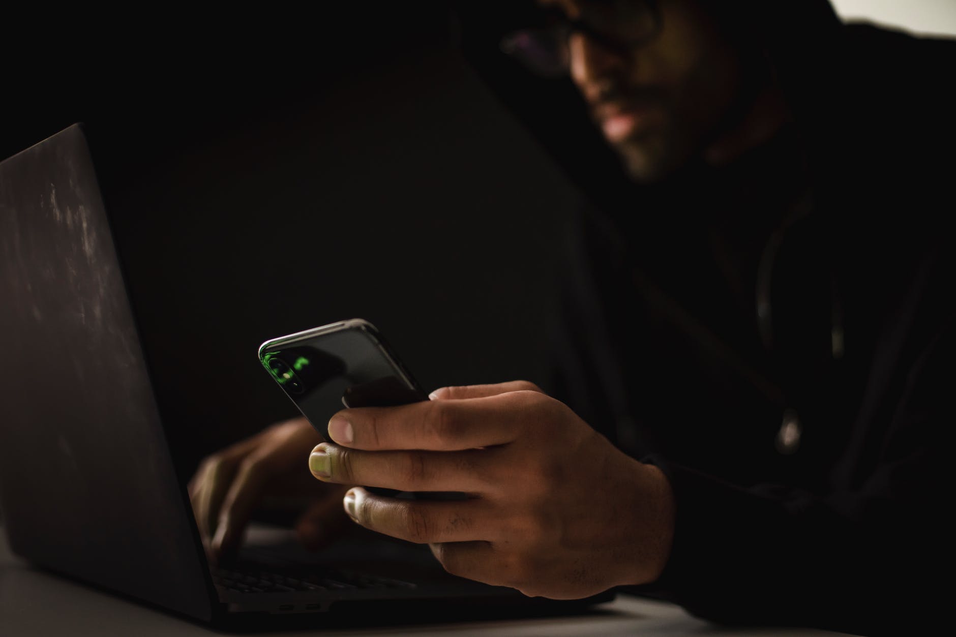 crop ethnic hacker with smartphone typing on laptop in dark room
